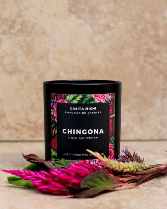 A black matte glass candle vessel with a floral pattern label that reads 'CHINGONA'. The candle is surrounded by decorative flowers and leaves.