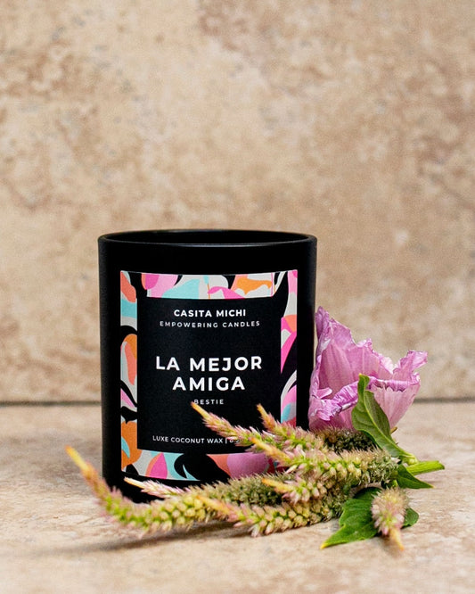An image of a black matte glass vessel candle with a label that reads 'LA MEJOR AMIGA' and the brand 'CASTA MICHI'. The candle is accompanied by decorative greenery.